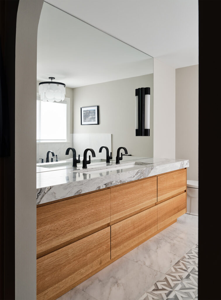 A modern bathroom vanity with a light wooden finish, white marble countertop, and black fixtures beneath a unique black and white wall sconce