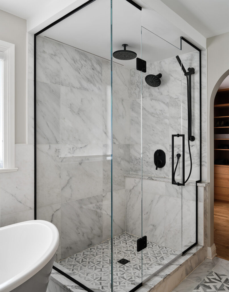 A sleek corner shower with marble walls, patterned tile floor, and black hardware, adjacent to a white freestanding bathtub