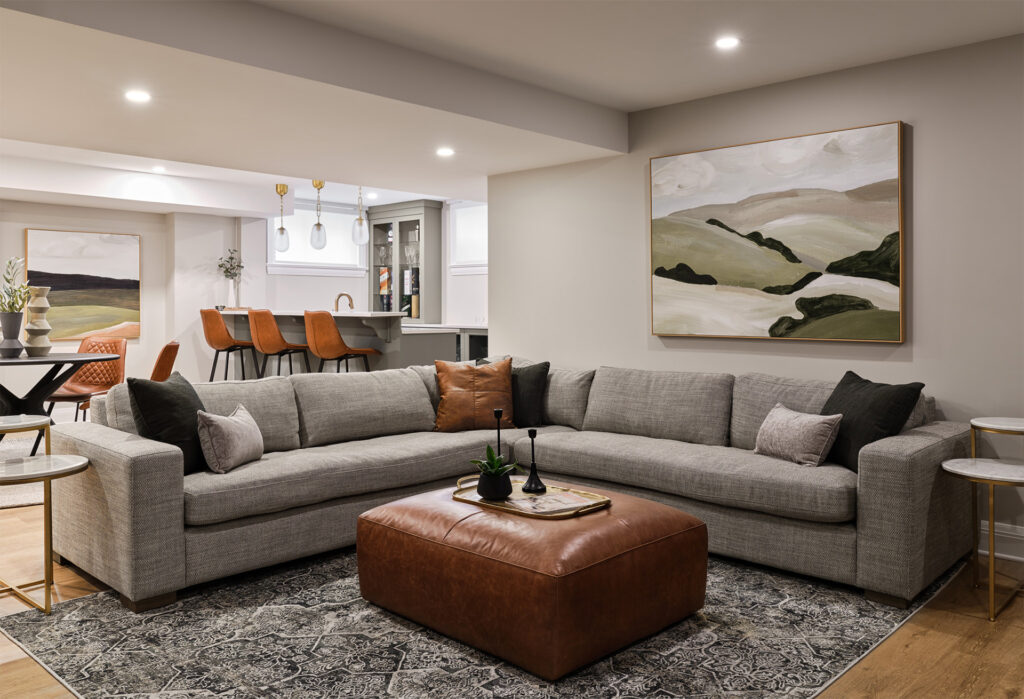 A modern living room with a sectional gray sofa, a large leather ottoman, an abstract painting, and an adjoining dining area