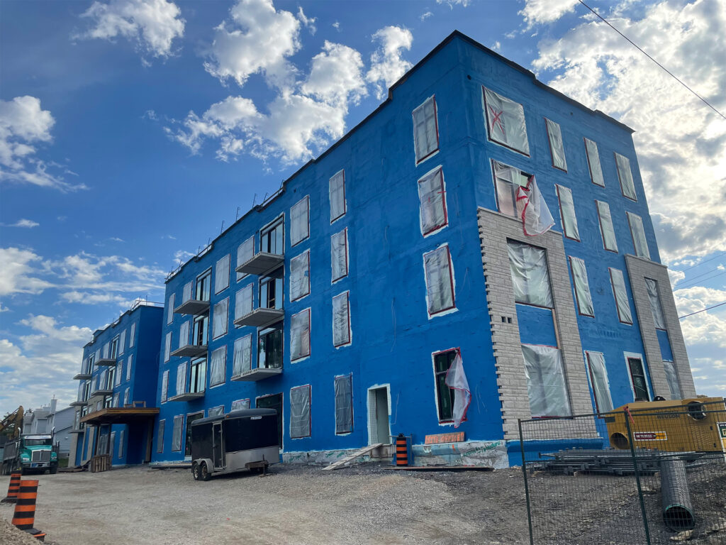 View of a multi-story building under construction with blue waterproofing material on its exterior walls, partially installed windows, and construction materials scattered around the site