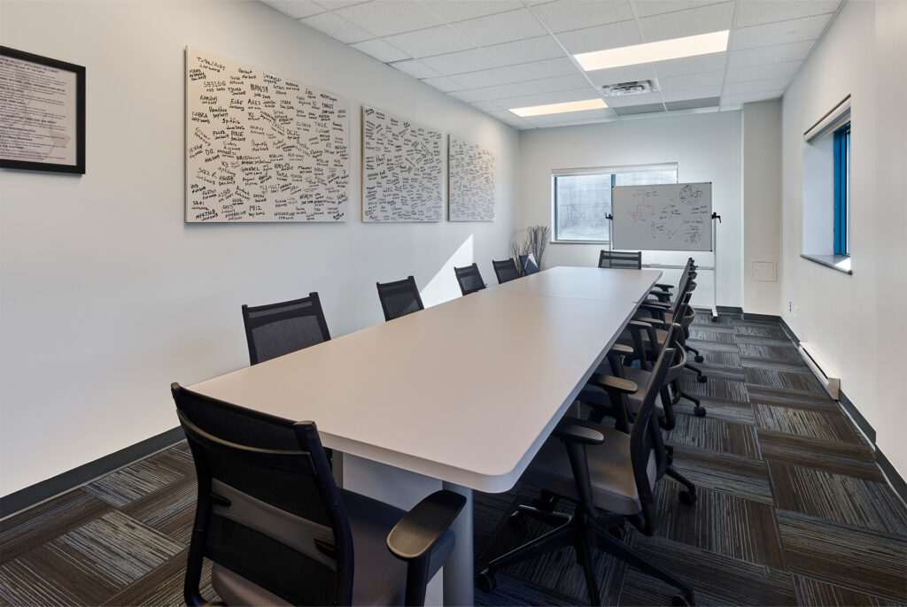 A modern meeting room with a large white conference table, black office chairs, carpeted floor, and a wall-mounted whiteboard