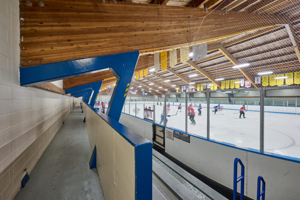 View from the spectator area of Fred Barrett Arena showing the ice rink and players through protective netting, with exposed wooden ceiling beams overhead