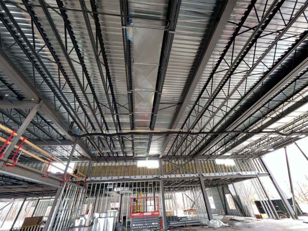 Interior of a large construction site with metal framing and open ceiling showing beams and ductwork, with construction lifts and materials on the ground