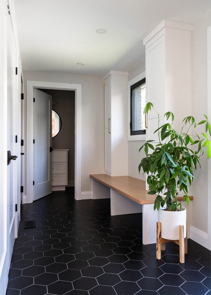 A modern hallway featuring geometric black floor tiles, white walls, a wooden bench, and a potted plant, creating a clean and inviting entryway