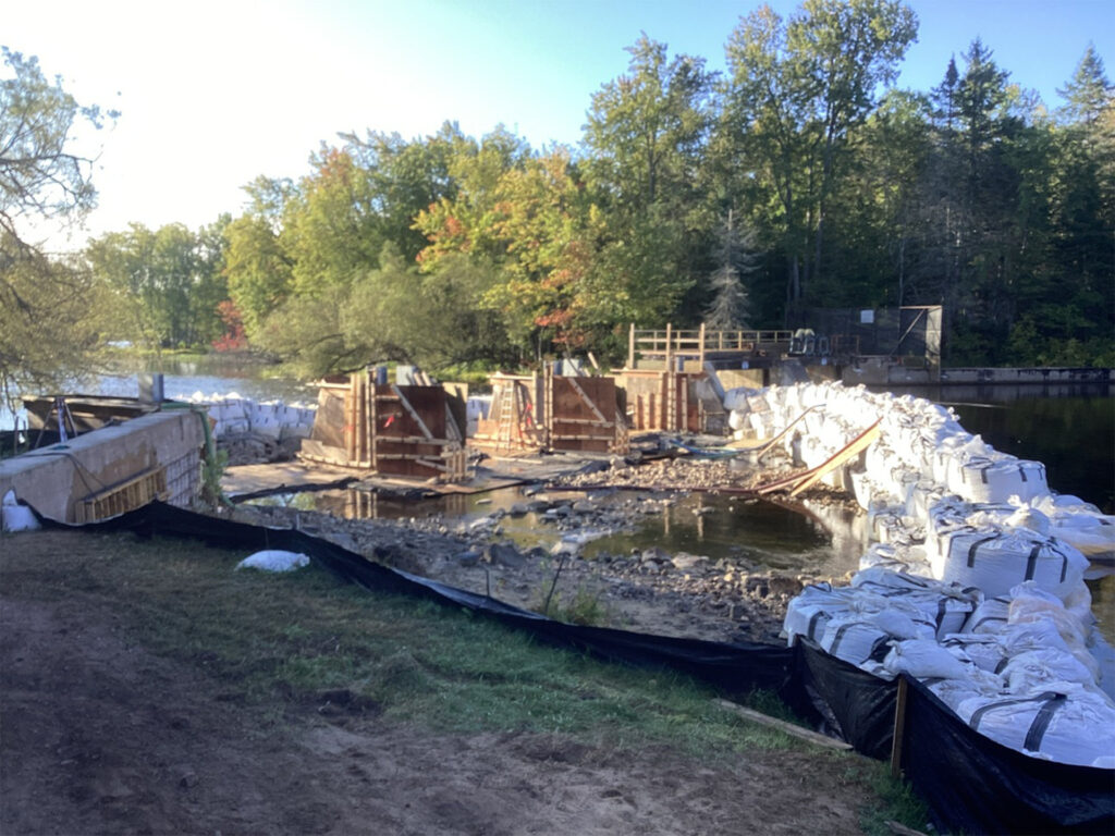 The image depicts a dam demolition site at Golden Lake, with partially dismantled structures, sandbag piles, and protective barriers in place