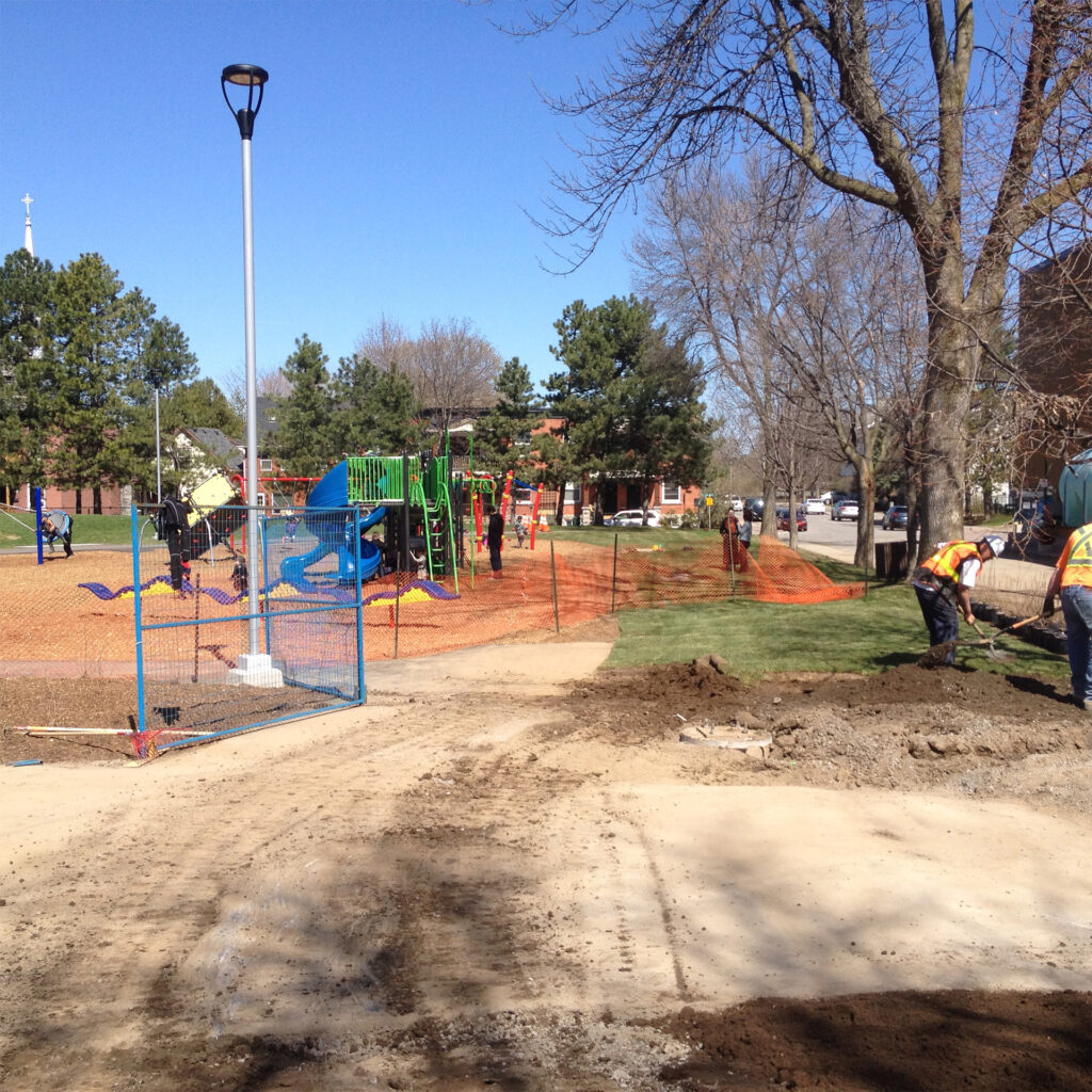 Park upgrade in progress with new playground equipment installed and workers landscaping the surrounding area