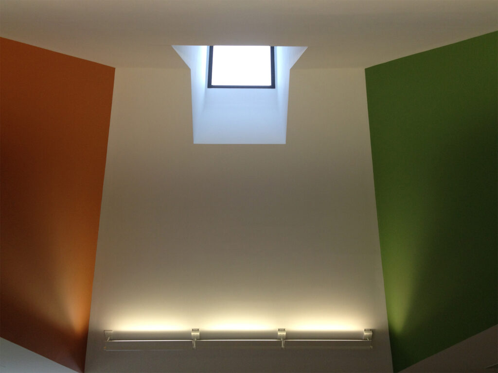 A room corner with a square skylight and walls painted in white, orange, and green, lit by a strip of wall-mounted lights