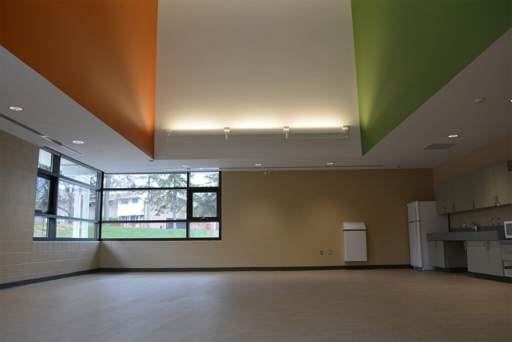 An empty room with a kitchenette, featuring a slanted ceiling with orange and green sections, track lighting, and large windows overlooking an outdoor area