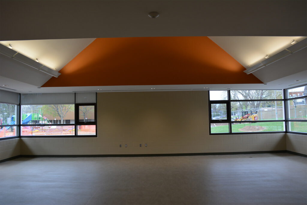 Interior view of a room with a high ceiling featuring an orange accent, track lighting, and large windows overlooking a construction area outside