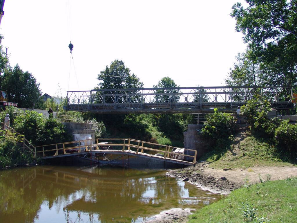 Installation of a new metal truss bridge over a river with construction equipment and temporary wooden supports in place