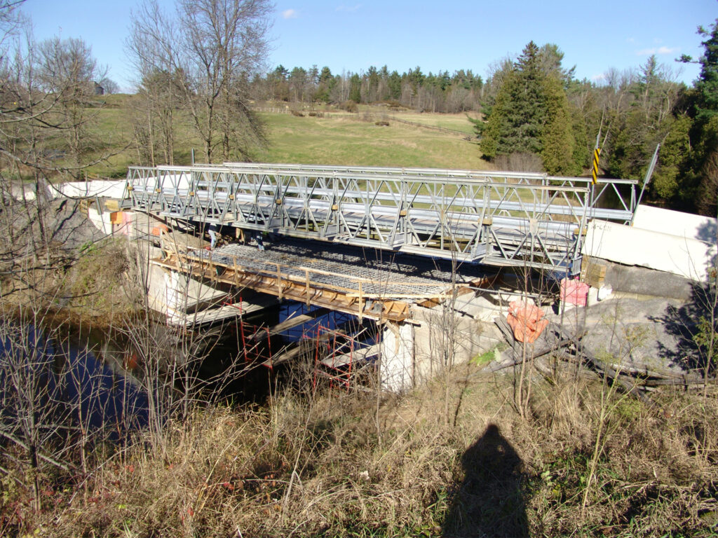 A metal truss bridge under construction with scaffolding and temporary support structures visible, set in a rural area.