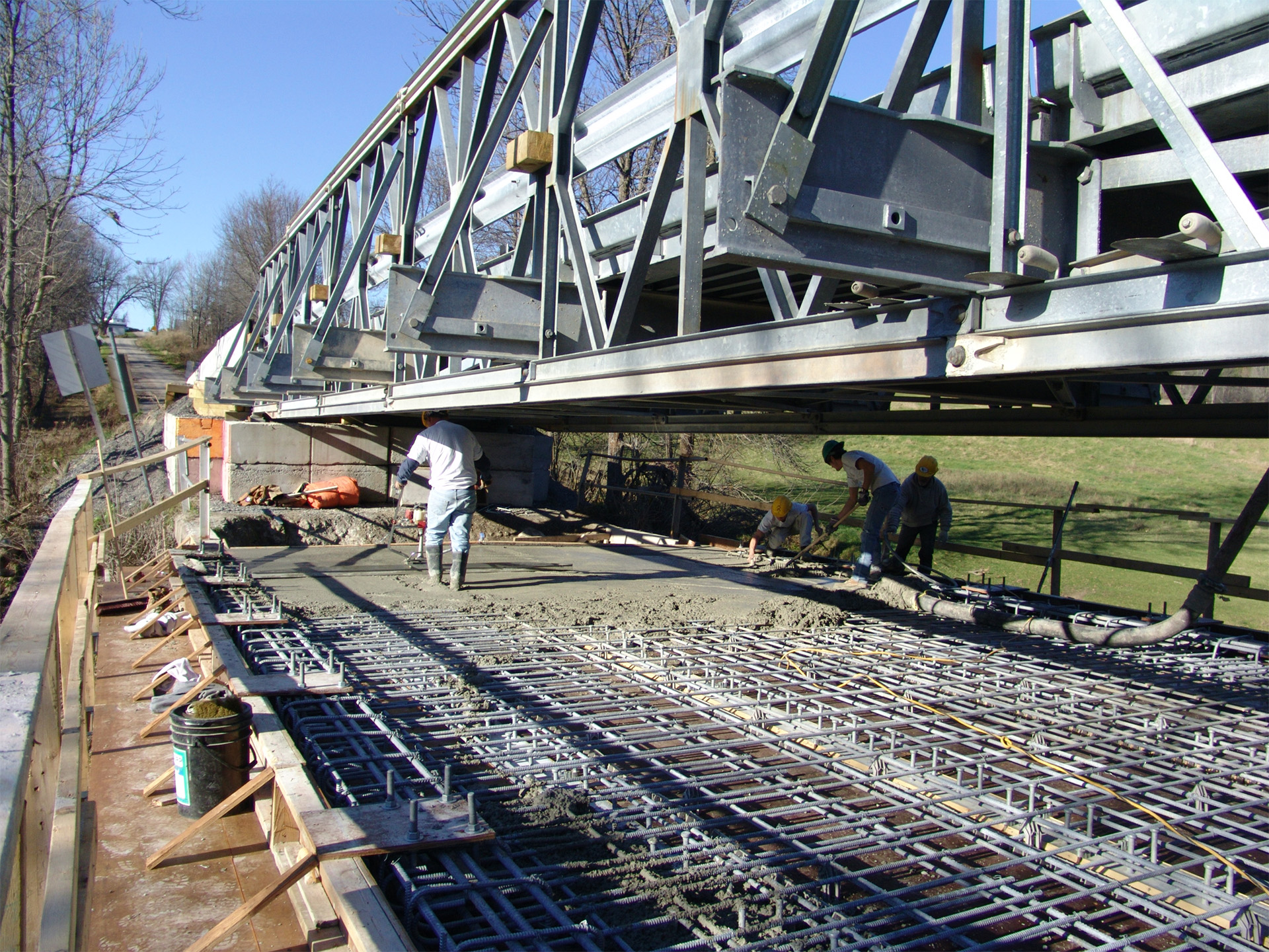 The image shows a construction site at Mississippi Mills where workers are actively engaged in laying concrete and reinforcing bars for a bridge under construction, with a metal truss bridge visible above them