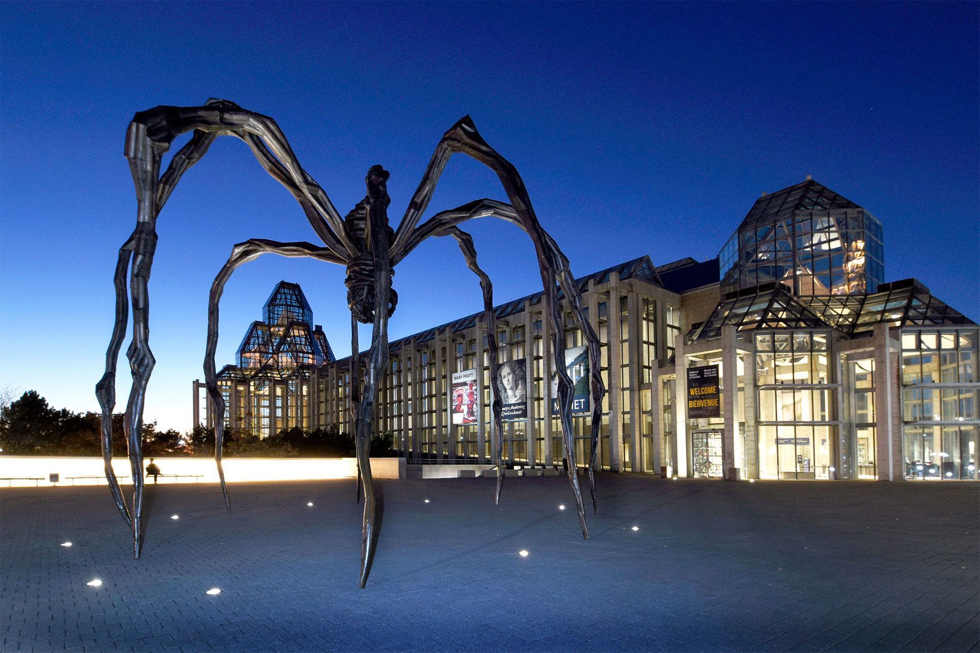The image features the exterior of the National Gallery of Canada at dusk with a large spider sculpture in the foreground and the gallery's modern glass and stone architecture illuminated in the background