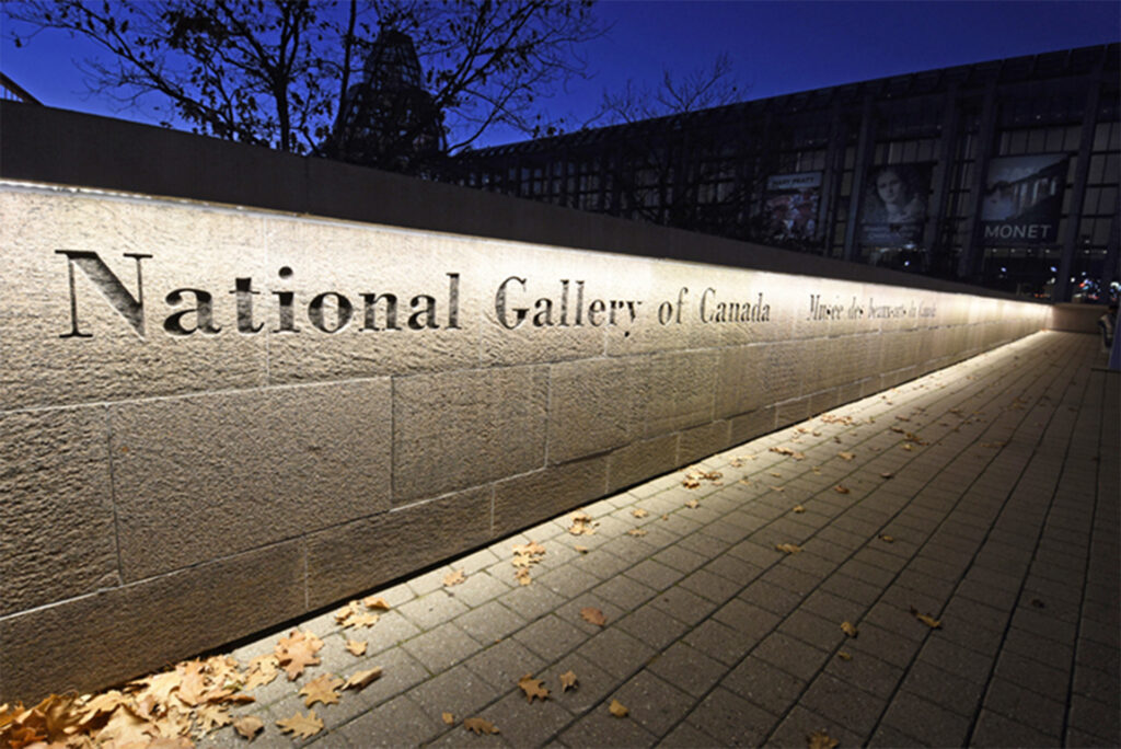 The image displays the engraved sign of the National Gallery of Canada on a stone wall, illuminated at night, with fallen leaves on the sidewalk and a background of the gallery's glass facade