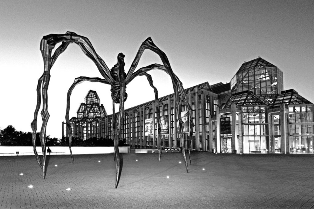 Monochrome image of a large spider sculpture in front of the National Gallery, with clear skies above and lights illuminating the plaza